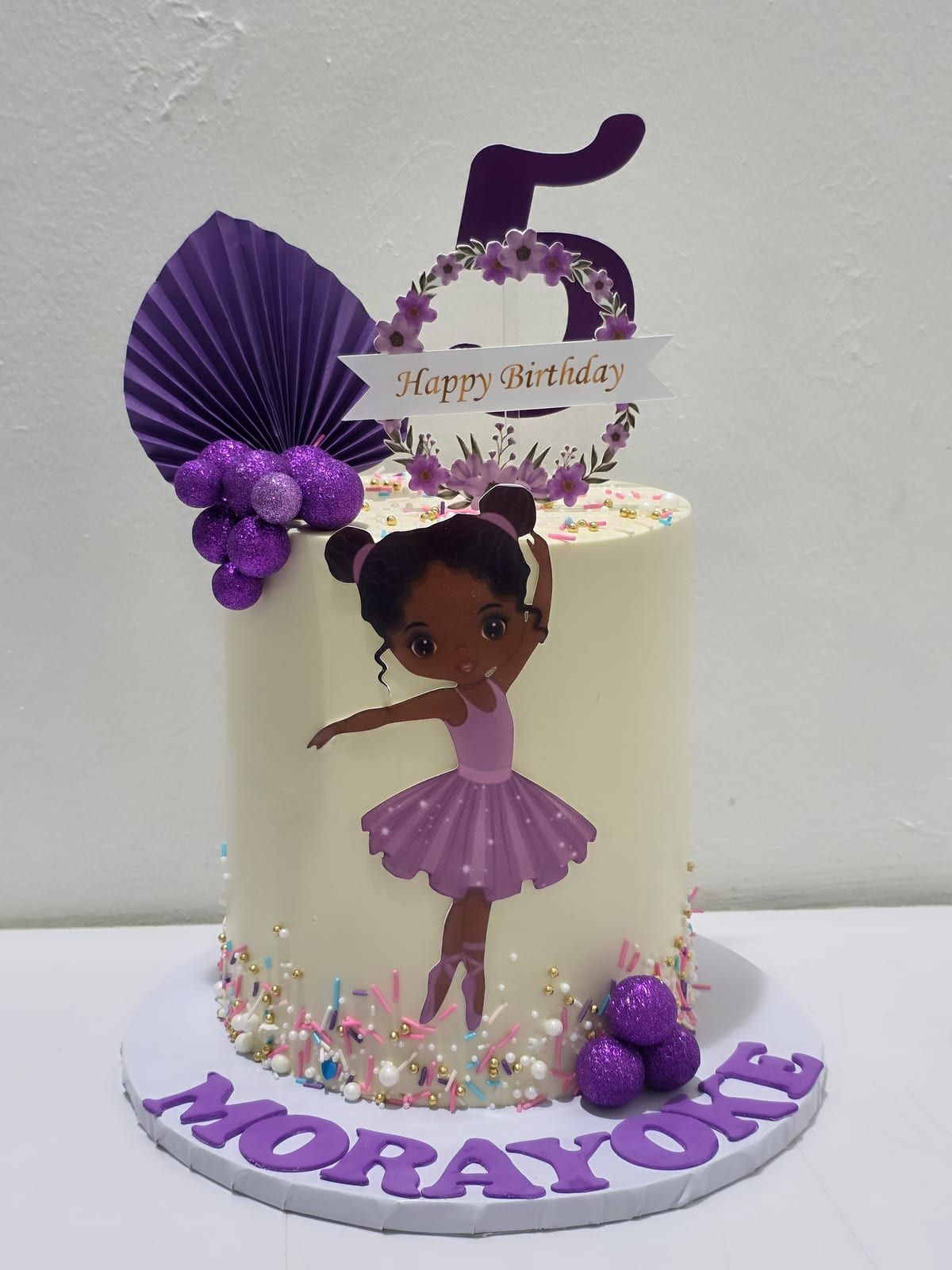 Tall ivory cake with a ballerina figure in a purple tutu, a purple fan topper, sugar pearls and violet sprinkles around the base