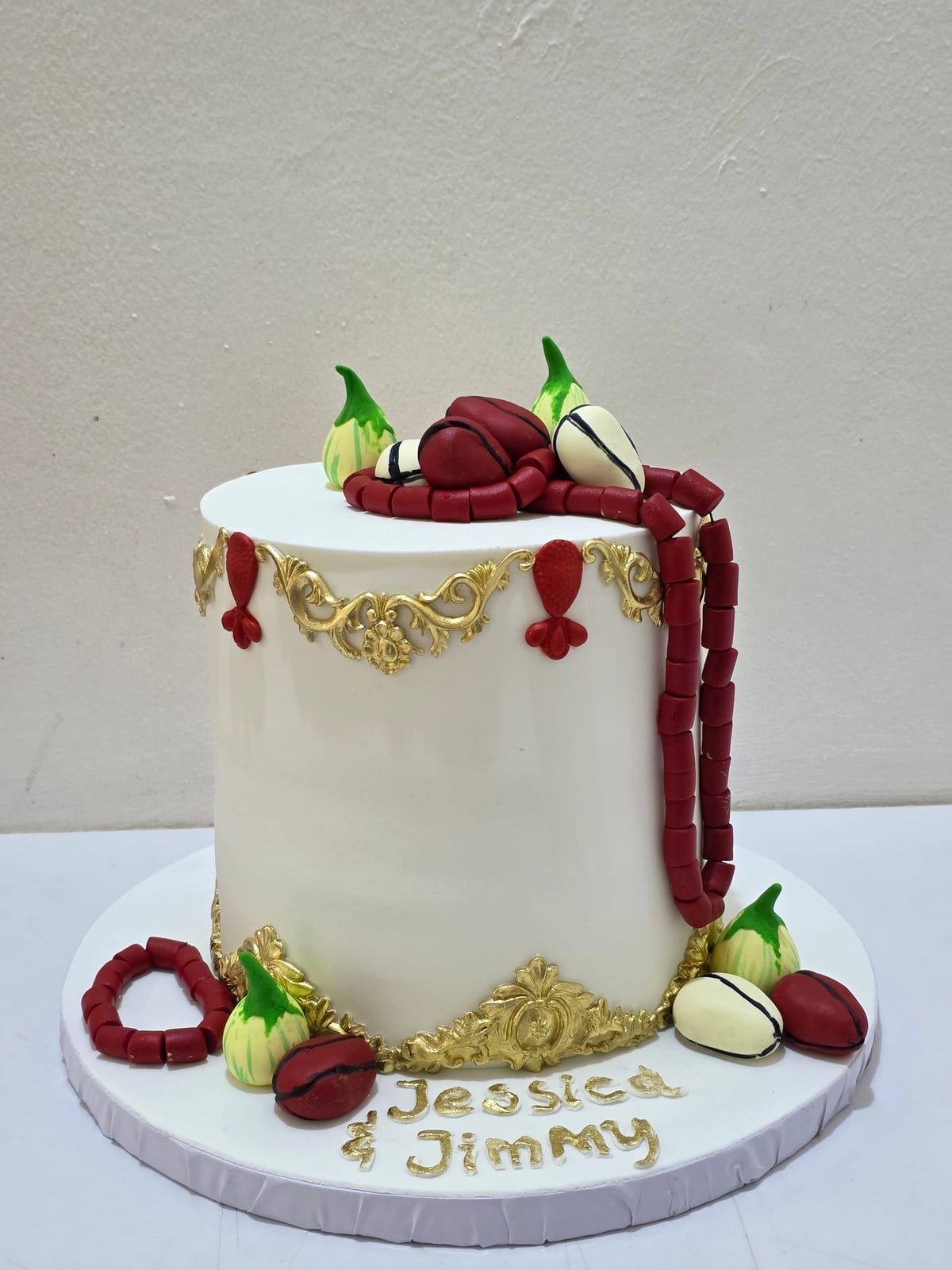 Ivory fondant cake trimmed with gold filigree, draped with strings of red coral beads, with garden eggs and kola nuts modelled in sugar around the top and base, and two names piped in gold