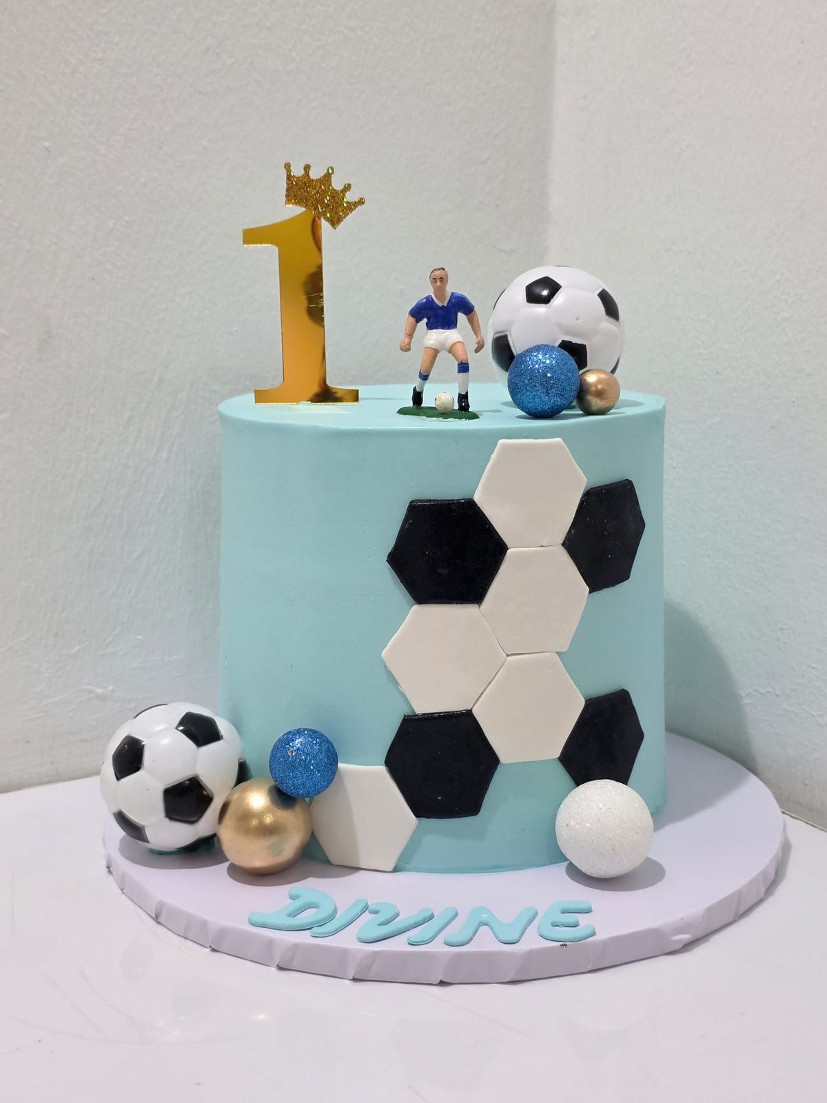 Tall pale-blue cake with black and white hexagon panels, a small footballer figure and a football on top, a gold "1" with a crown, and the name piped in blue at the base