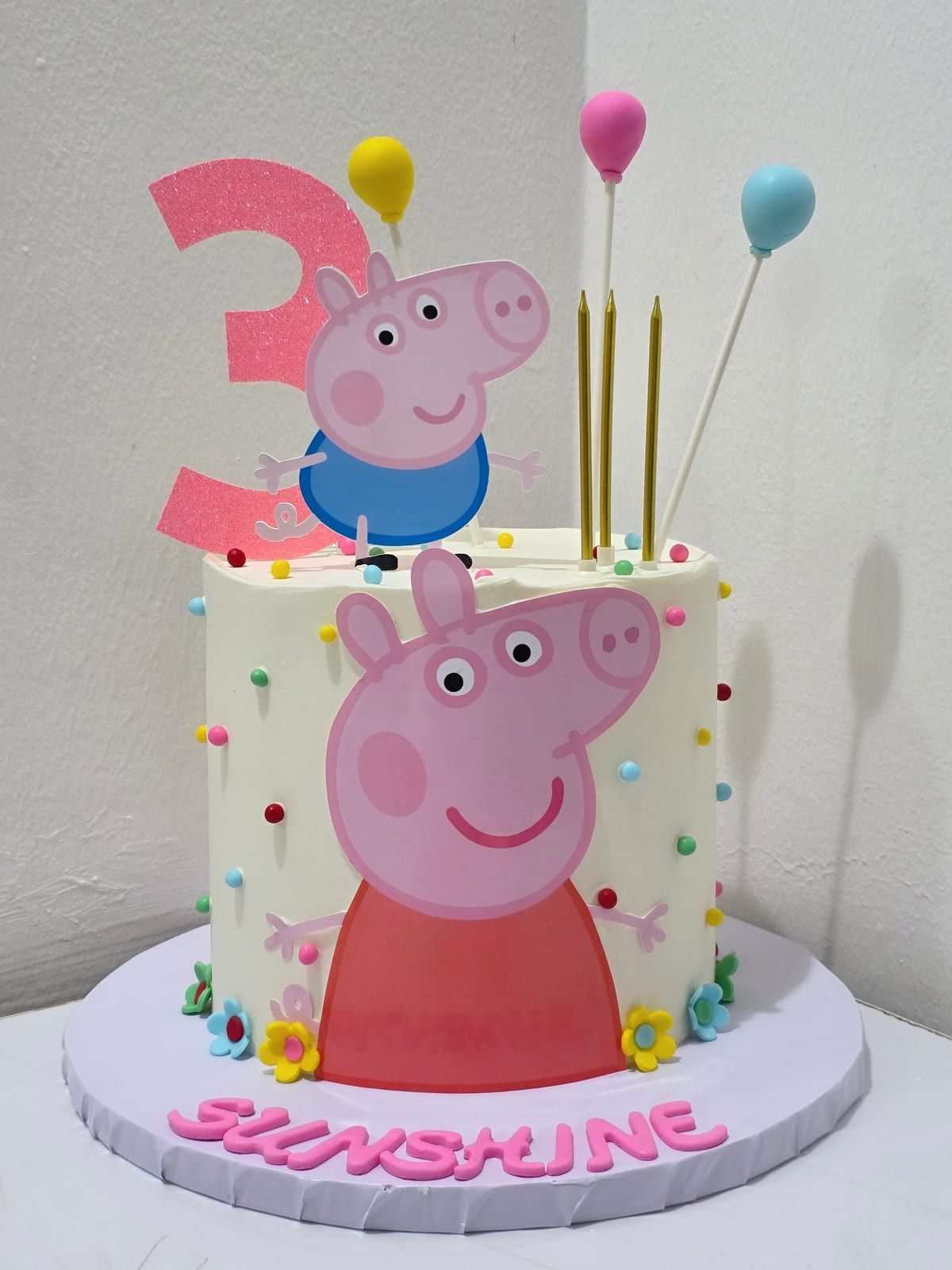 Tall ivory cake covered in coloured sprinkles, with a large cartoon pig on the front, a pink glitter "3", balloon toppers and the name piped in pink at the base