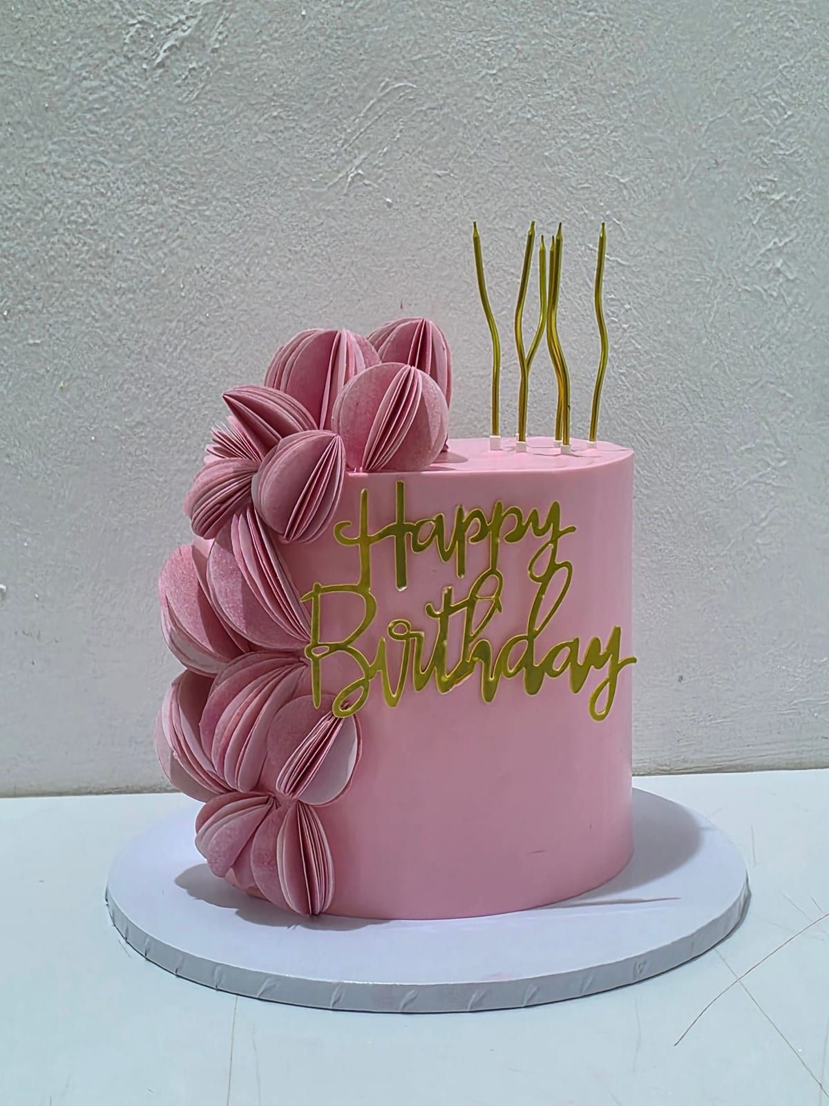 Tall pink cake with a fan of piped ruffles sweeping down one side, gold script topper and slim gold candles
