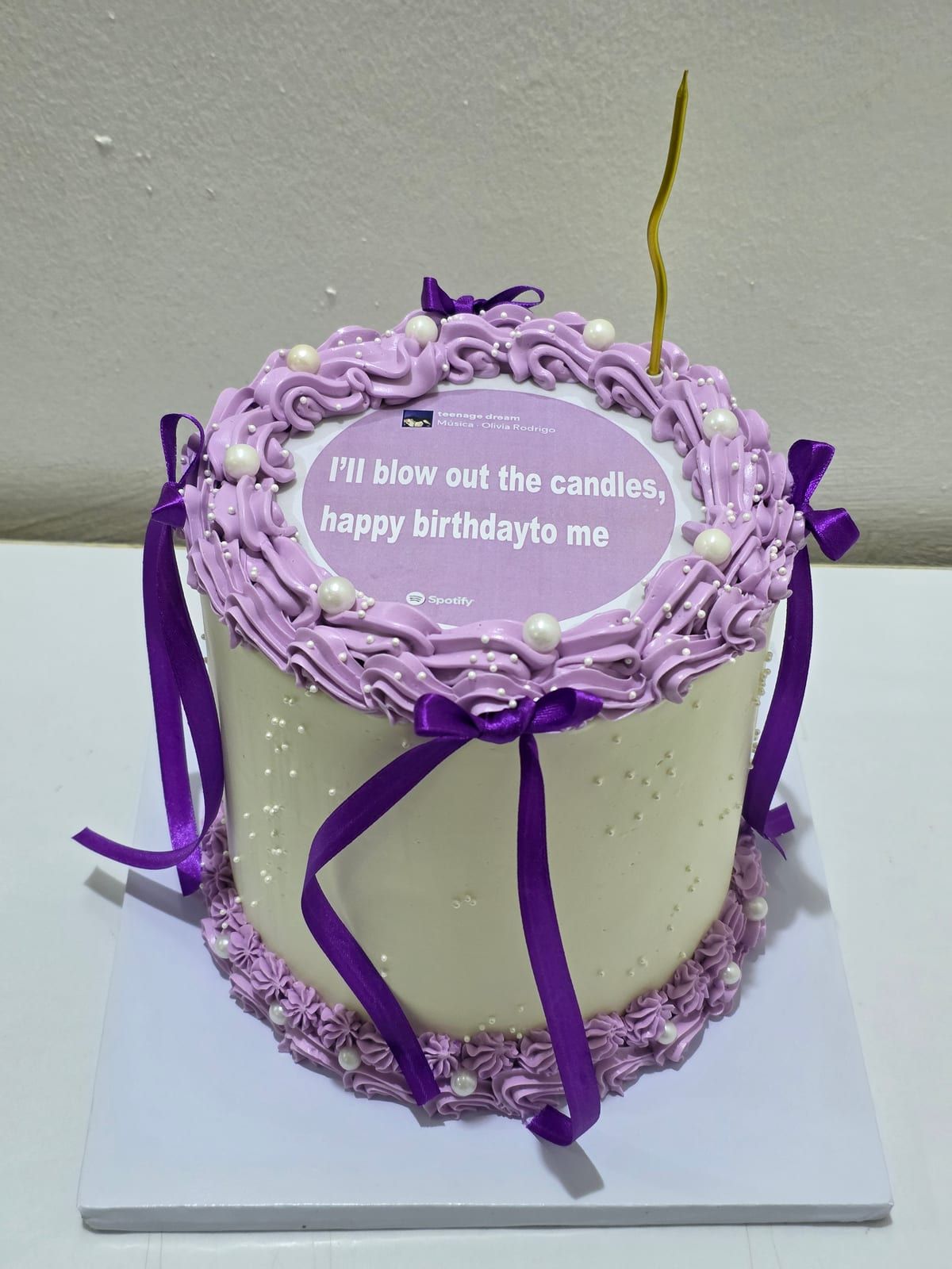 Tall ivory cake with lilac vintage-style piping and pearls at the top and base, purple ribbons down the sides, a printed lyric card on top and a single gold candle
