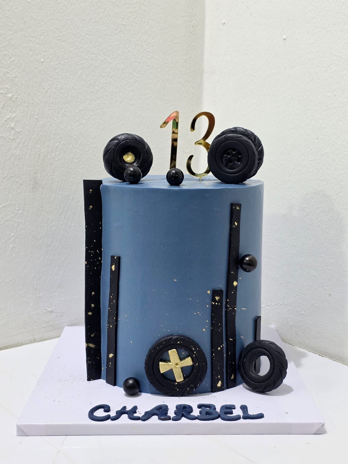 Tall steel-blue cake flecked with gold, with black sugar tyres and bars around the sides, a gold cross emblem, more tyres and a gold "13" on top, and the name piped in blue on the board