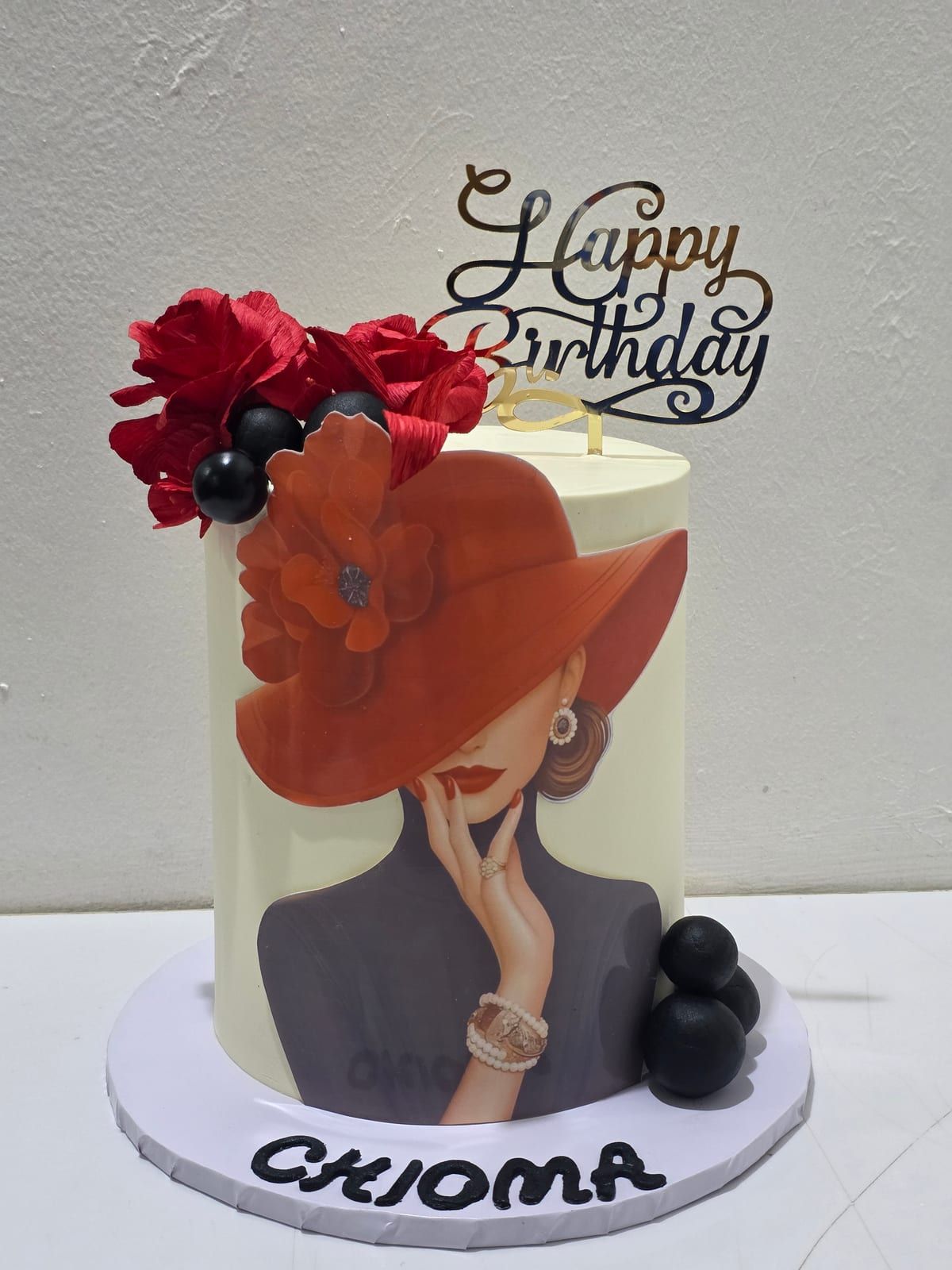 Tall ivory cake with an edible print of a woman in a wide red hat, deep-red sugar flowers and black spheres on top, a black "Happy Birthday" topper, and the name piped in black around the base