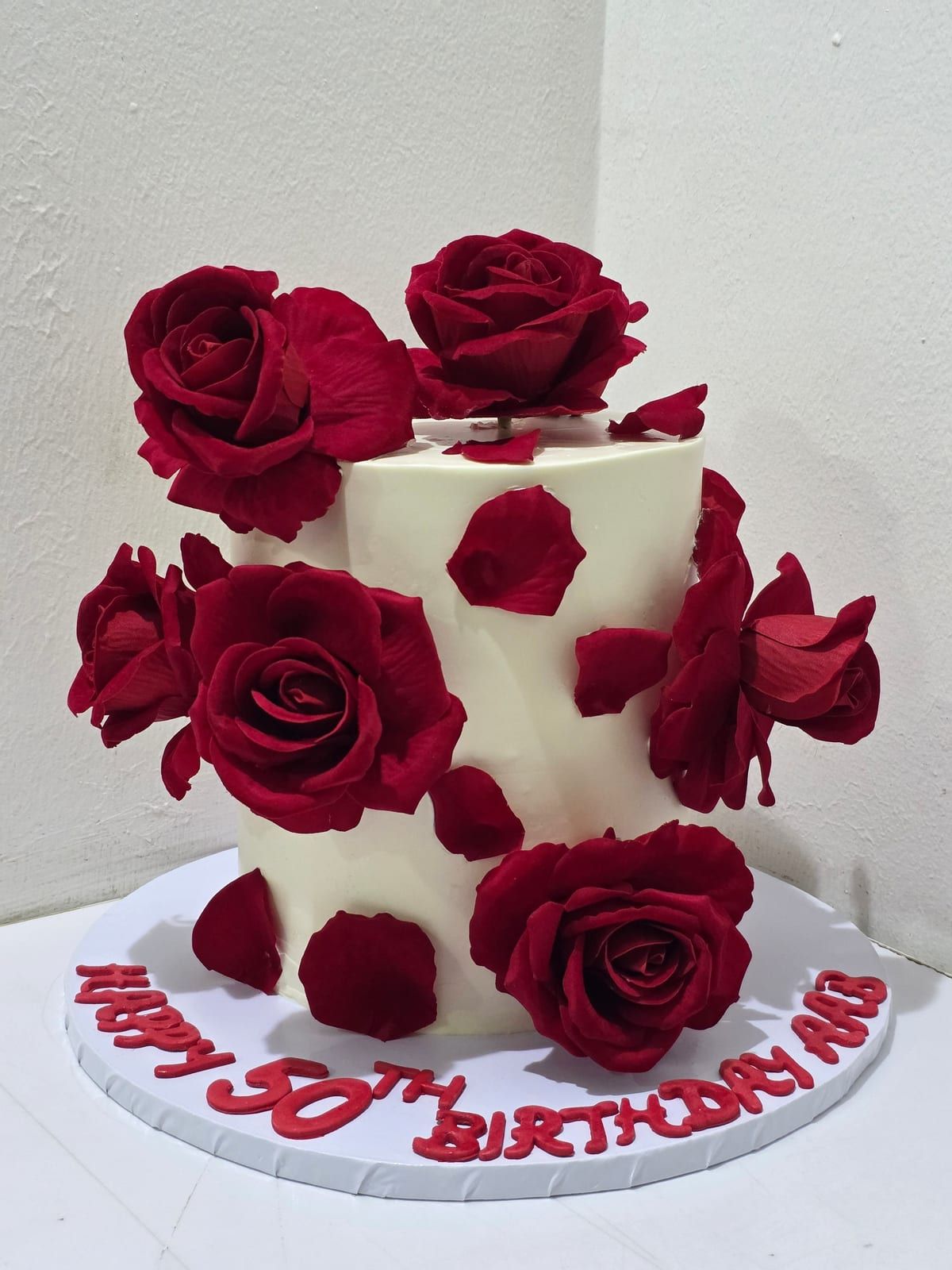 Tall ivory cake covered in large deep-red sugar roses and scattered petals, with red lettering piped around the base