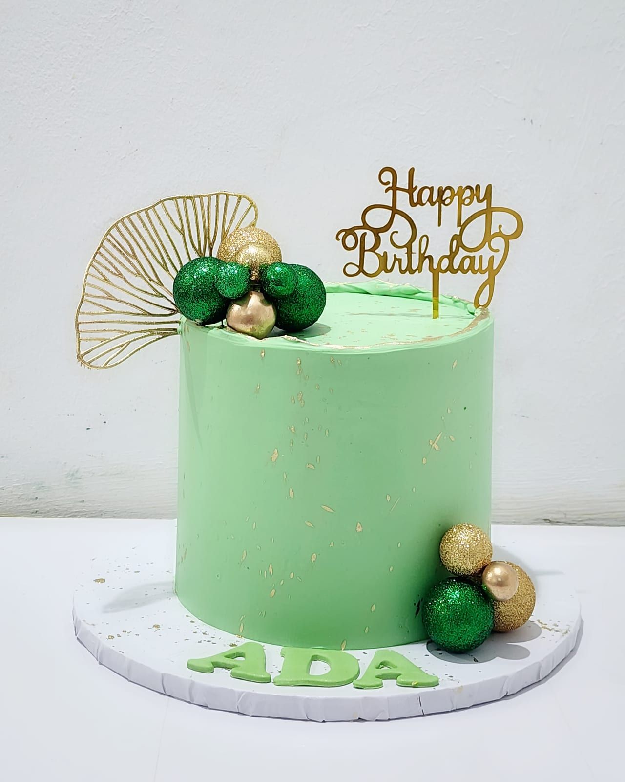 Tall mint-green cake flecked with gold, with a gold leaf fan, green and gold spheres, a gold "Happy Birthday" topper and the name piped in green at the base