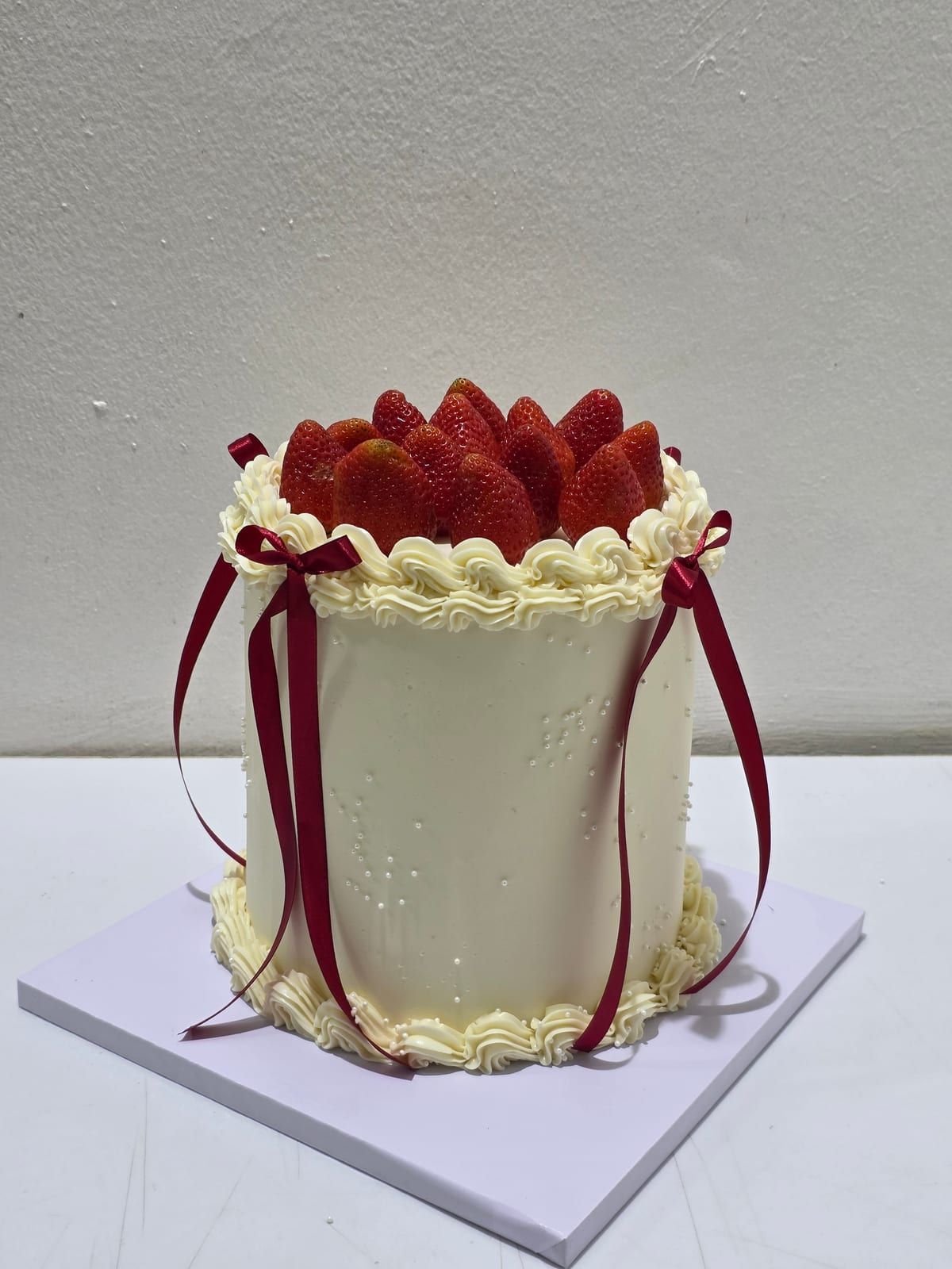 Tall ivory cake with piped cream shells at the top and base, a pile of fresh strawberries on top, and burgundy ribbons tied down the sides
