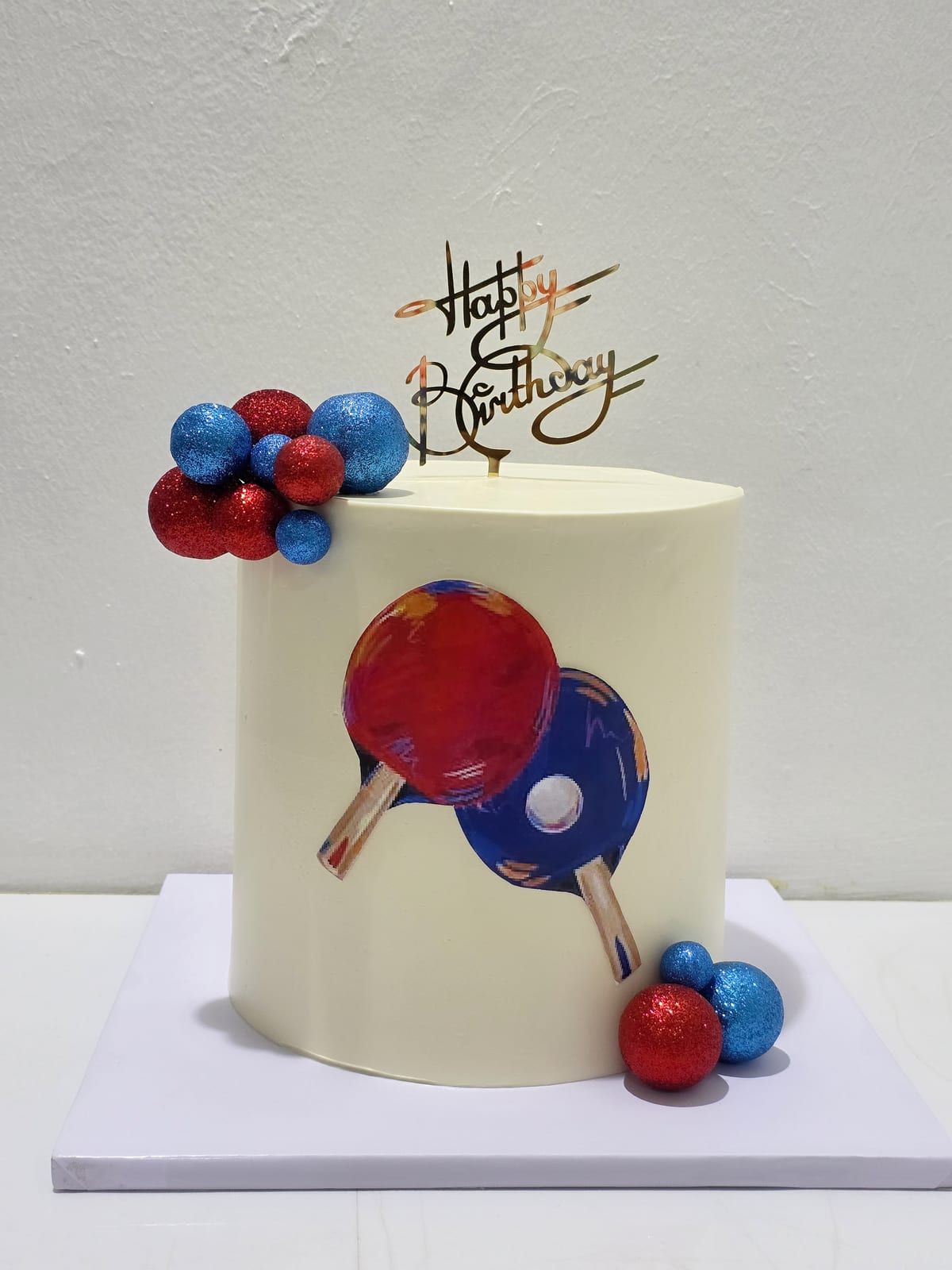 Tall ivory cake decorated with two modelled table-tennis bats, red and blue balls and clustered spheres