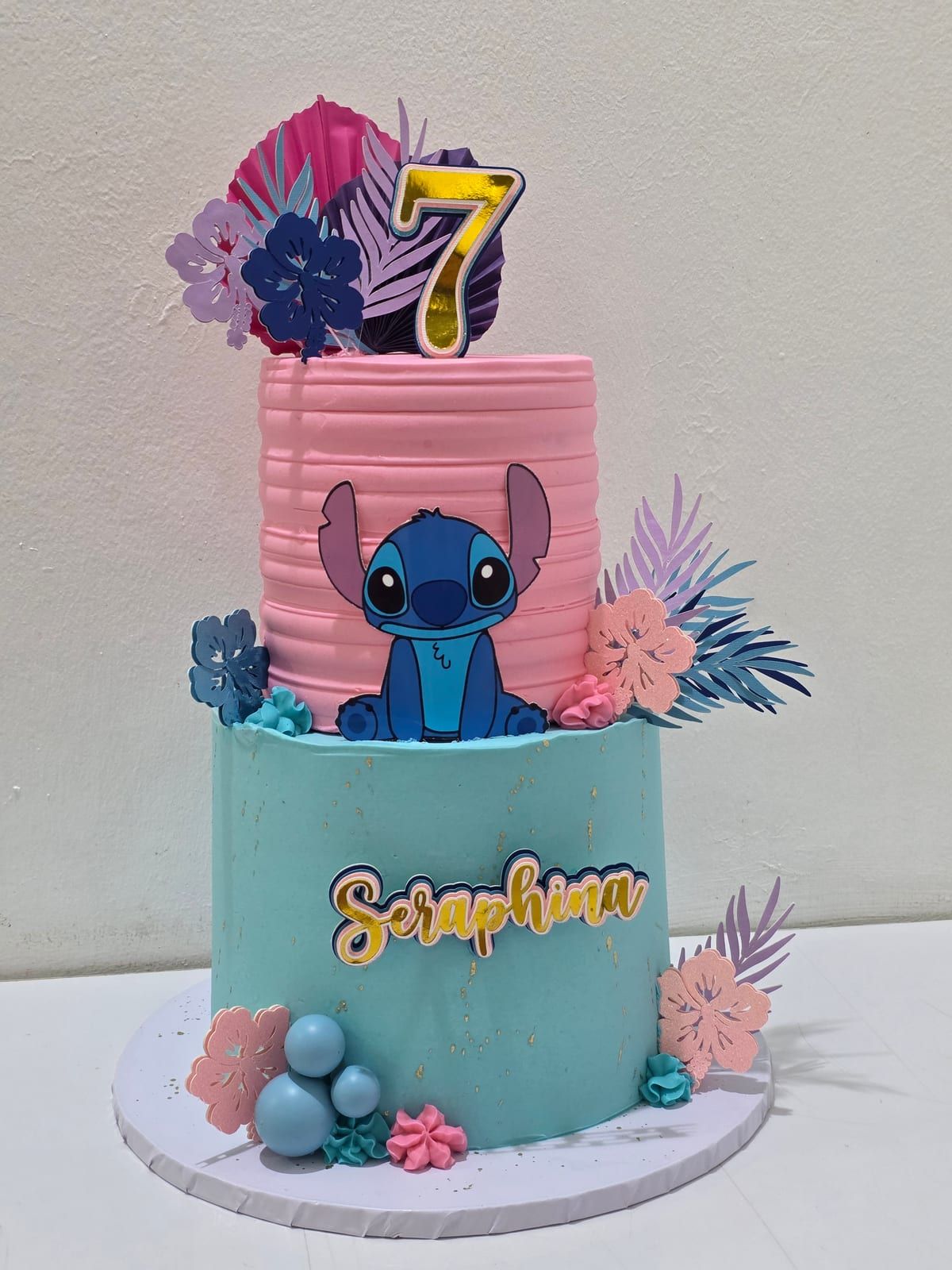 Two-tier cake with a teal buttercream base and a pink ruffled upper tier, topped with a character figure, sugar flowers and pastel spheres