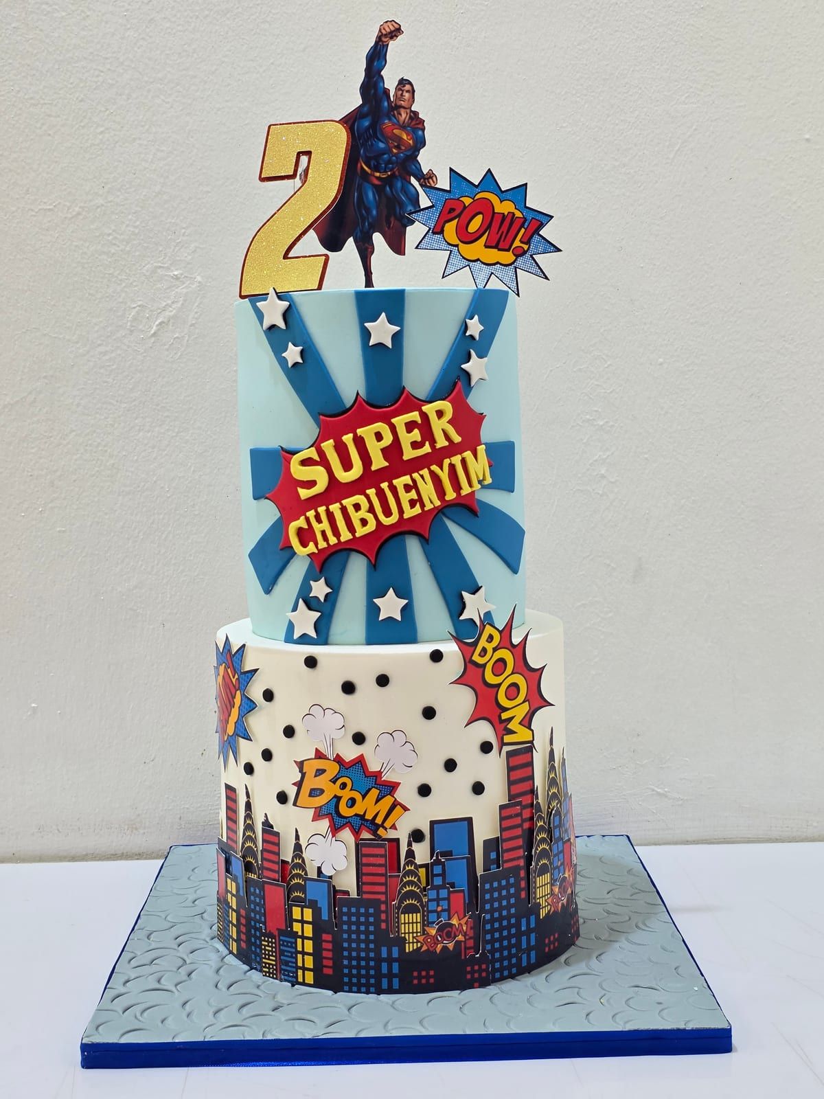 Two-tier fondant cake with a comic-book theme — a blue starburst upper tier with a superhero figure, and a lower tier decorated with a red and blue city skyline