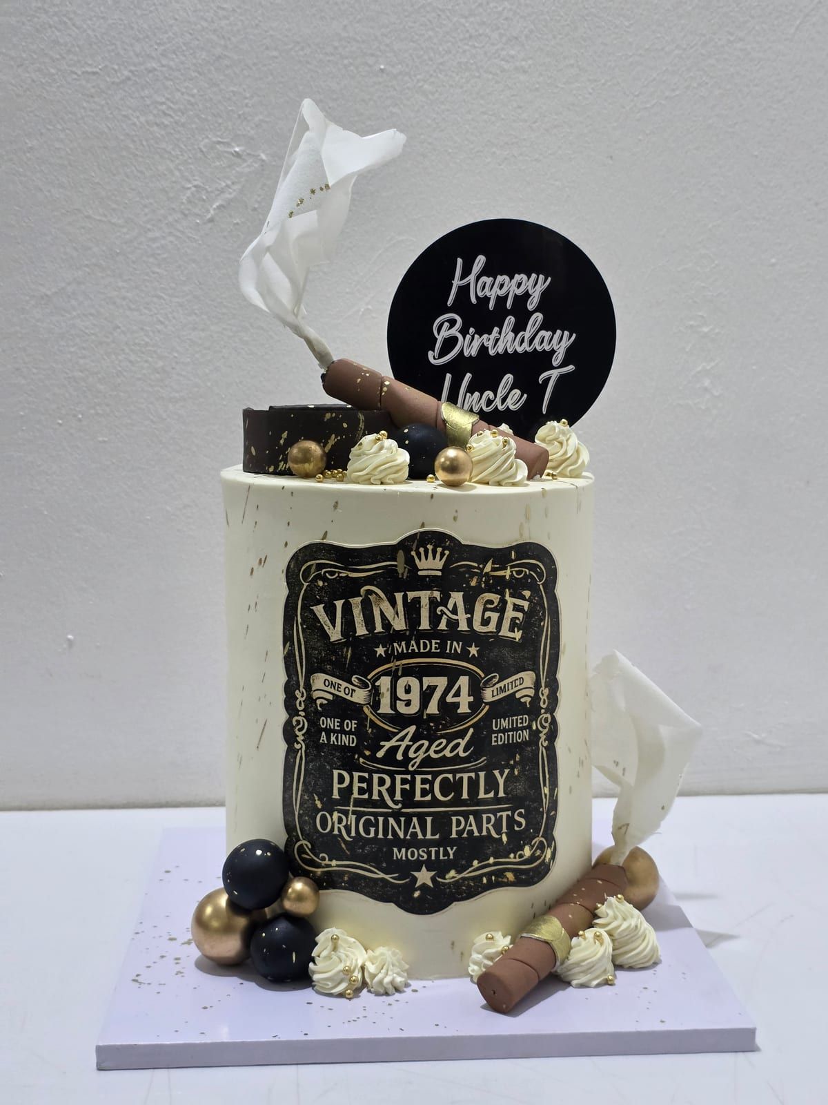 Tall ivory cake with a black "Vintage 1974 — Aged Perfectly" label, cigar and whisky-glass toppers, black and gold spheres, and a round black "Happy Birthday" topper